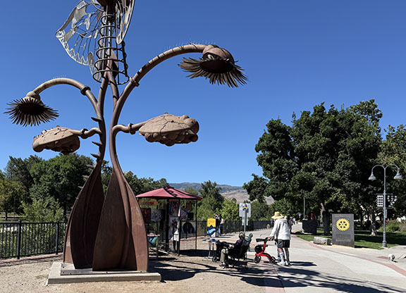 large metal sculpture on riverwalk with an art display under an umbrella with people on sidewalk
